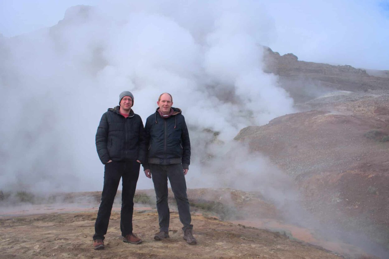 Zwei Männer stehen vor einem dampfenden Geysir.