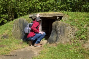 Eine Person mit Rucksack und Hut hockt am Eingang eines kleinen, mit Steinen ausgekleideten unterirdischen Bauwerks, das von Gras und Vegetation umgeben ist.