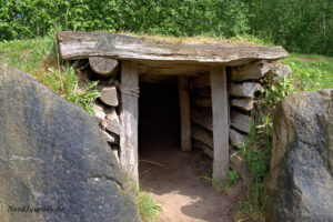 Ein kleines hölzernes Eingangsgebäude, flankiert von großen Steinen und teilweise mit Gras bedeckt, führt in einen Tunnel oder Unterstand in einer natürlichen, grünen Umgebung.