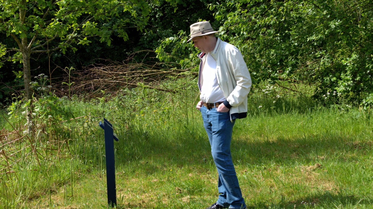Ein Mann mit Hut, weißer Jacke und Jeans steht mit den Händen in den Taschen da und betrachtet ein Informationsschild auf einer Rasenfläche mit Bäumen im Hintergrund.