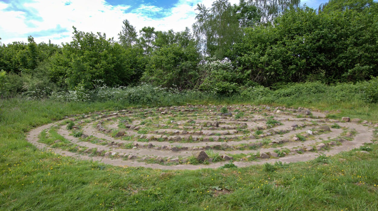 Ein kreisförmiges Steinlabyrinth befindet sich auf einem Grasgelände, umgeben von grünem Laub und Bäumen unter einem teilweise bewölkten Himmel.