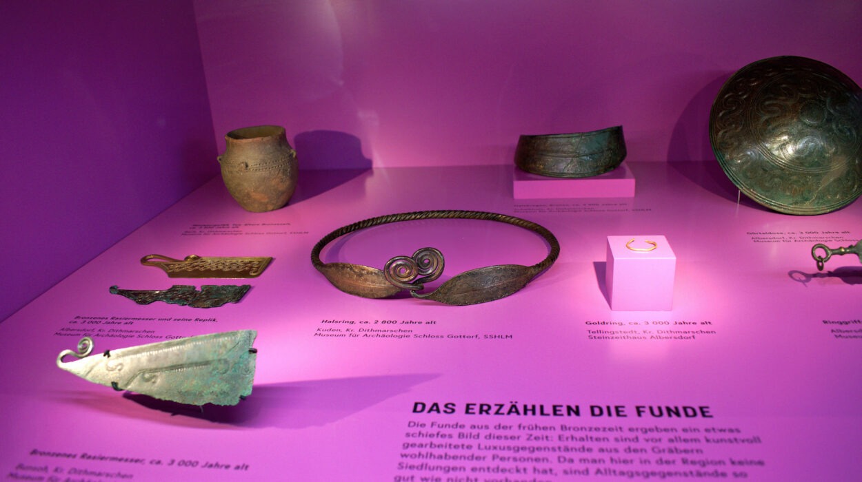 Eine Museumsausstellung mit verschiedenen Bronzeartefakten, darunter ein Armband, eine Brosche und andere Gegenstände, vor einem rosa Hintergrund mit beschreibenden Beschriftungen auf Deutsch.