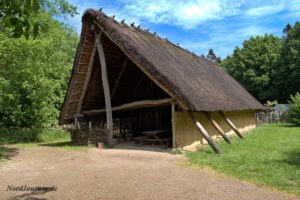 Ein Gebäude mit Holz- und Strohdach steht auf einer von Bäumen umgebenen Rasenfläche unter einem klaren blauen Himmel.