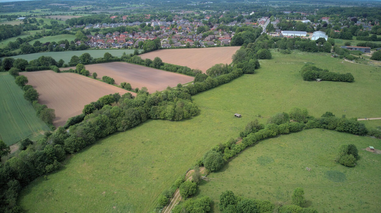 Luftaufnahme einer ländlichen Landschaft mit grünen Feldern, Baumgruppen und einem kleinen Dorf in der Ferne unter einem teilweise bewölkten Himmel.