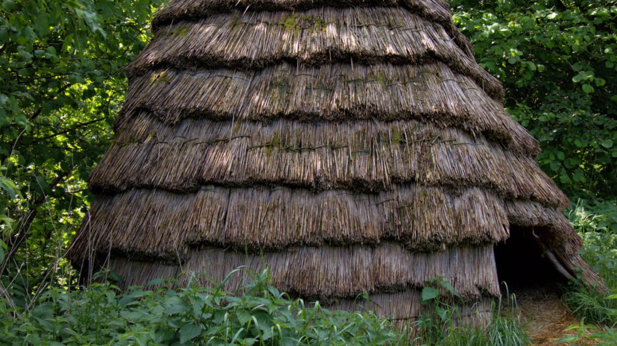 Eine kleine, runde, strohgedeckte Hütte mit kegelförmigem Dach ist von dichtem grünem Laub umgeben. Ein schmaler Feldweg führt zum Eingang.