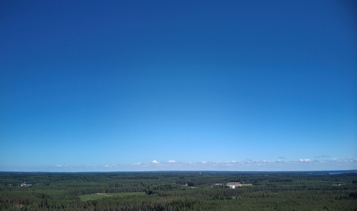 Luftaufnahme einer ausgedehnten Waldlandschaft mit verstreuten Seen unter einem klaren blauen Himmel. Im Hintergrund sind der Horizont und entfernte Wolken zu sehen.