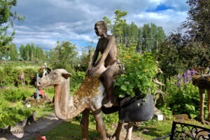 In einem Garten steht die Statue einer Person, die auf einem Kamel reitet. Auf dem Rücken des Kamels wachsen Pflanzen aus Behältern. Im Hintergrund sieht man üppiges Grün und einen teilweise bewölkten Himmel.