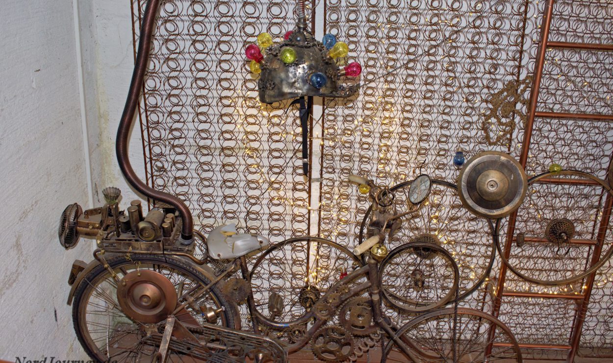 Ein Kunstwerk im Steampunk-Stil zeigt ein Fahrrad aus Zahnrädern, Federn und Rohren vor einem Hintergrund aus Metallspulen.