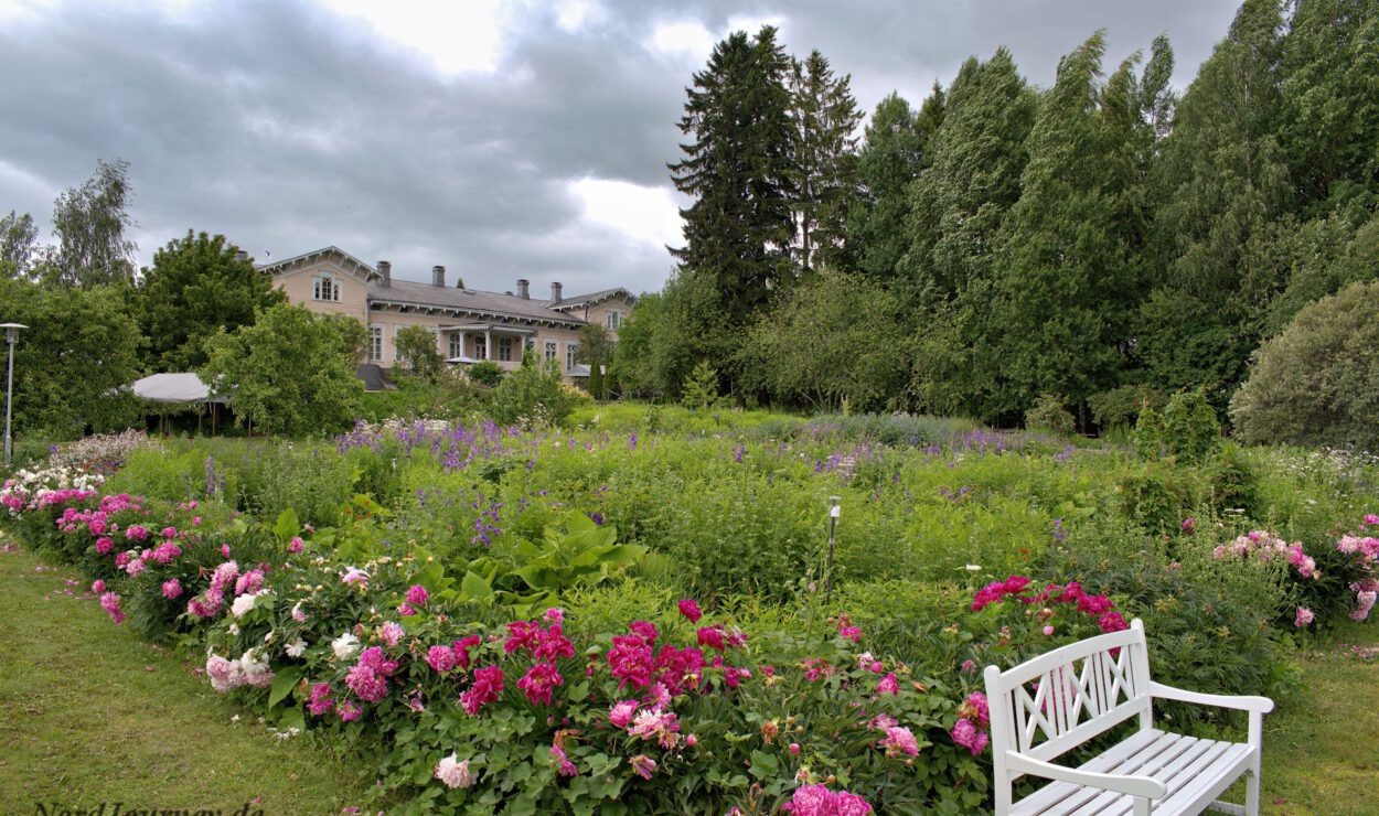 Ein Garten mit bunten Blumen, einer weißen Bank auf der rechten Seite und einem großen Haus im Hintergrund unter einem bewölkten Himmel.