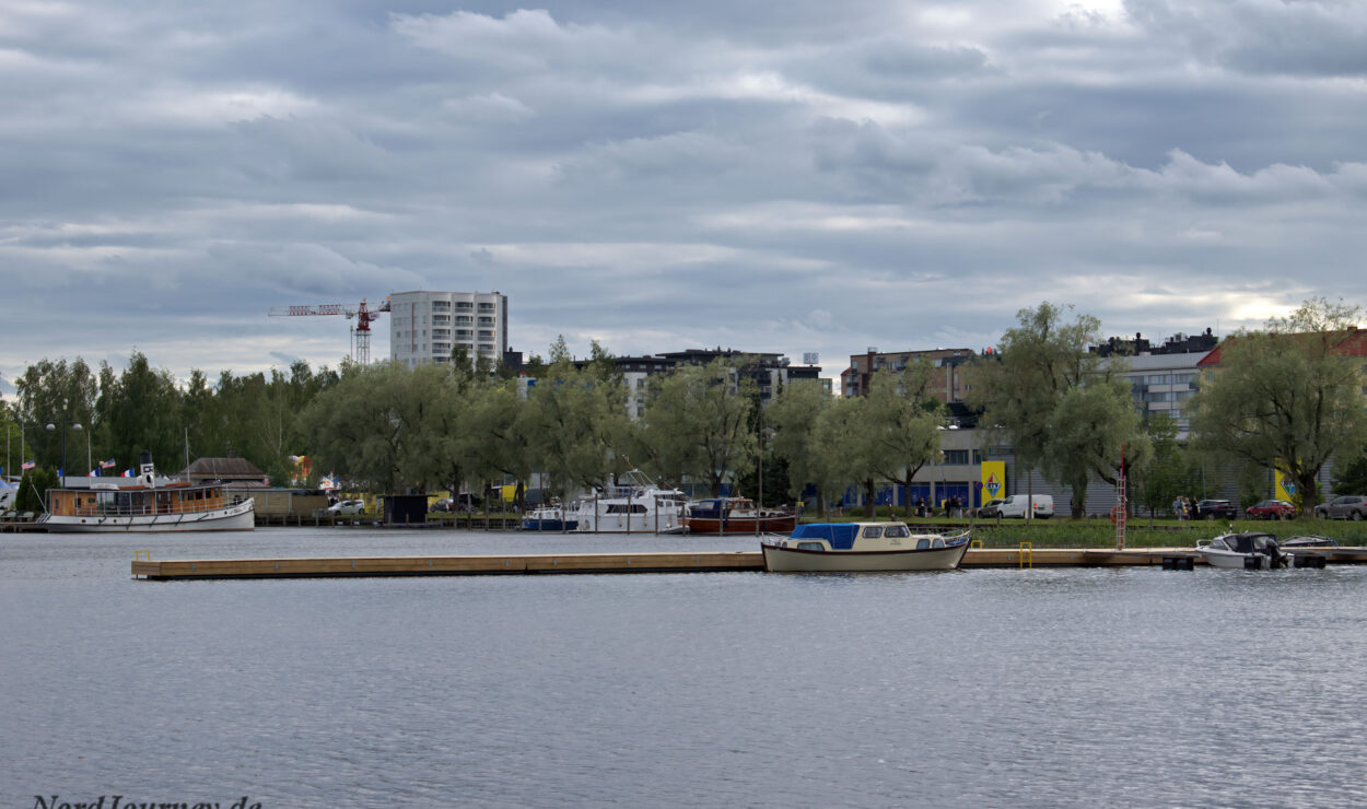 Eine Szene am Wasser mit mehreren Booten, die an einem Ufer angedockt sind. Im Hintergrund sind Bäume und Gebäude unter einem bewölkten Himmel zu sehen.