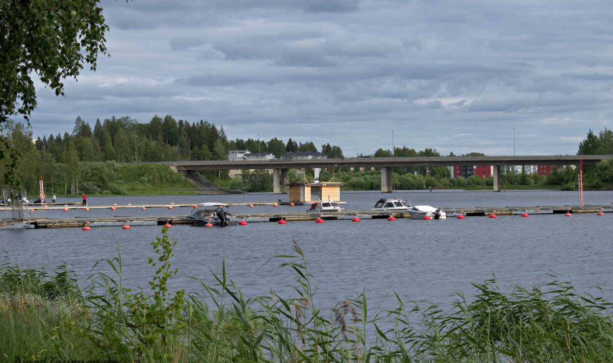 Eine Flussszene mit einer Reihe schwimmender Docks, mehreren festgemachten Booten und einer Brücke im Hintergrund unter einem bewölkten Himmel. Im Vordergrund und entlang des Flussufers ist Vegetation zu sehen.