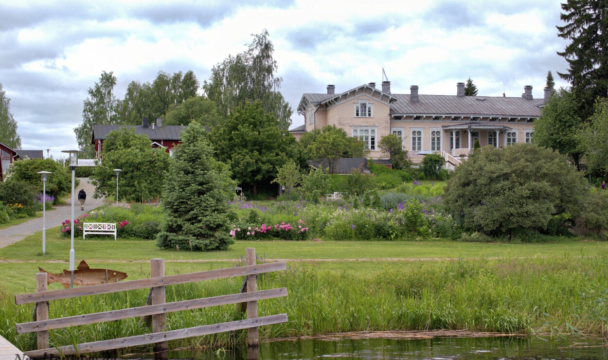 Hinter einem Holzzaun und einem Teich steht ein großes Haus mit gepflegtem Garten, umgeben von Grün und Bäumen unter einem bewölkten Himmel.