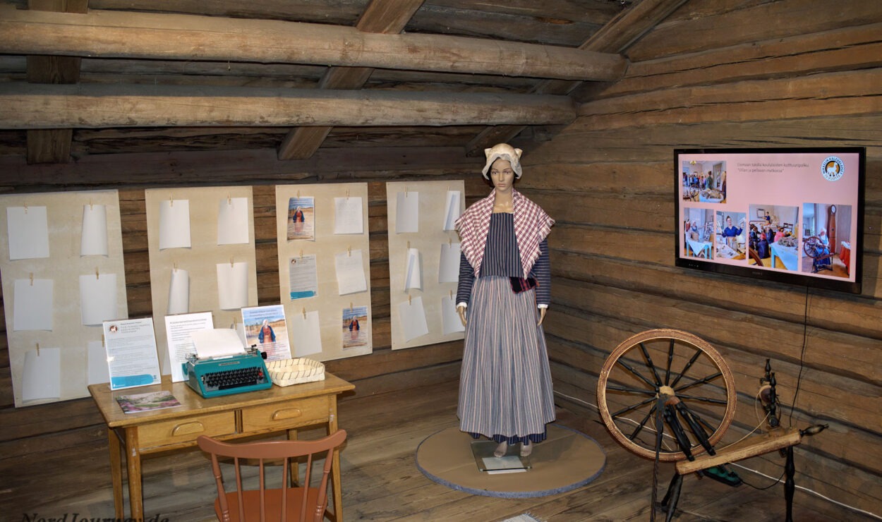 Eine historische Ausstellung zeigt eine Schaufensterpuppe in traditioneller Kleidung, ein Spinnrad, eine Schreibmaschine auf einem Holzschreibtisch, ausgestellte Dokumente und einen Fernsehbildschirm in einem rustikalen Holzraum.