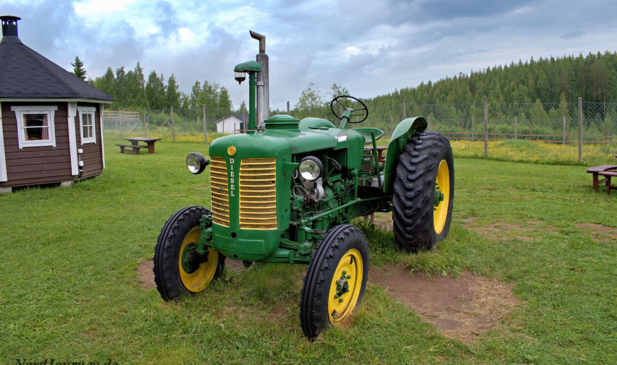 Ein alter grüner Traktor mit gelben Rädern steht auf einer Wiese neben einer Holzhütte, im Hintergrund sind ein Maschendrahtzaun und ein Wald zu sehen.