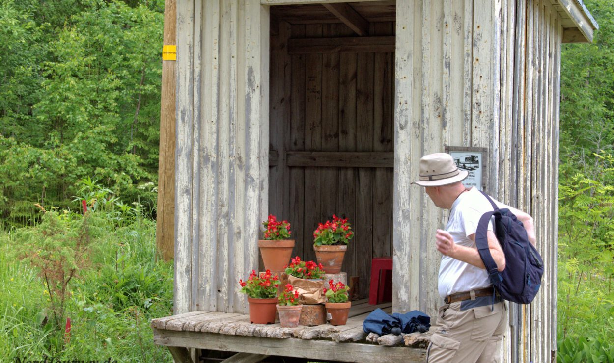 Ein Mann mit einem Rucksack steht vor einem kleinen Holzschuppen mit mehreren Töpfen roter Blumen auf einer Veranda, umgeben von Grün.