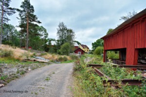 Unbefestigte Straße, die zu einer ländlichen Gegend mit roten und gelben Häusern führt, umgeben von Bäumen und einem roten Holzgebäude auf der rechten Seite. Bewölkter Himmel darüber.