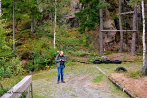 Ein Mann steht mit einer Kamera in der Hand auf einem Schotterweg in einem Waldstück. In der Nähe sind Reifen und Bäume gestapelt, im Hintergrund Felsen.