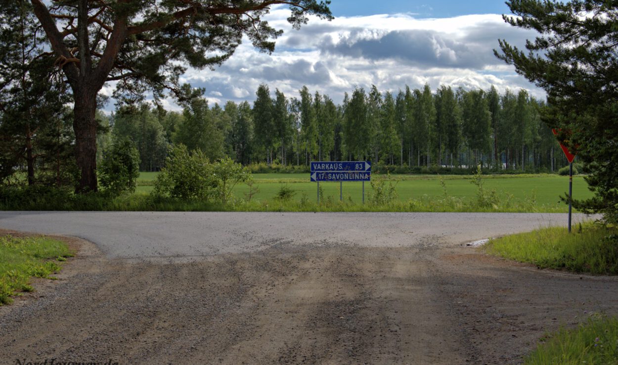 Eine ländliche Kreuzung mit einer Schotterstraße, die eine asphaltierte Straße kreuzt, umgeben von Bäumen. Ein blaues Wegweiser zeigt nach Varkaus und Savonlinna.