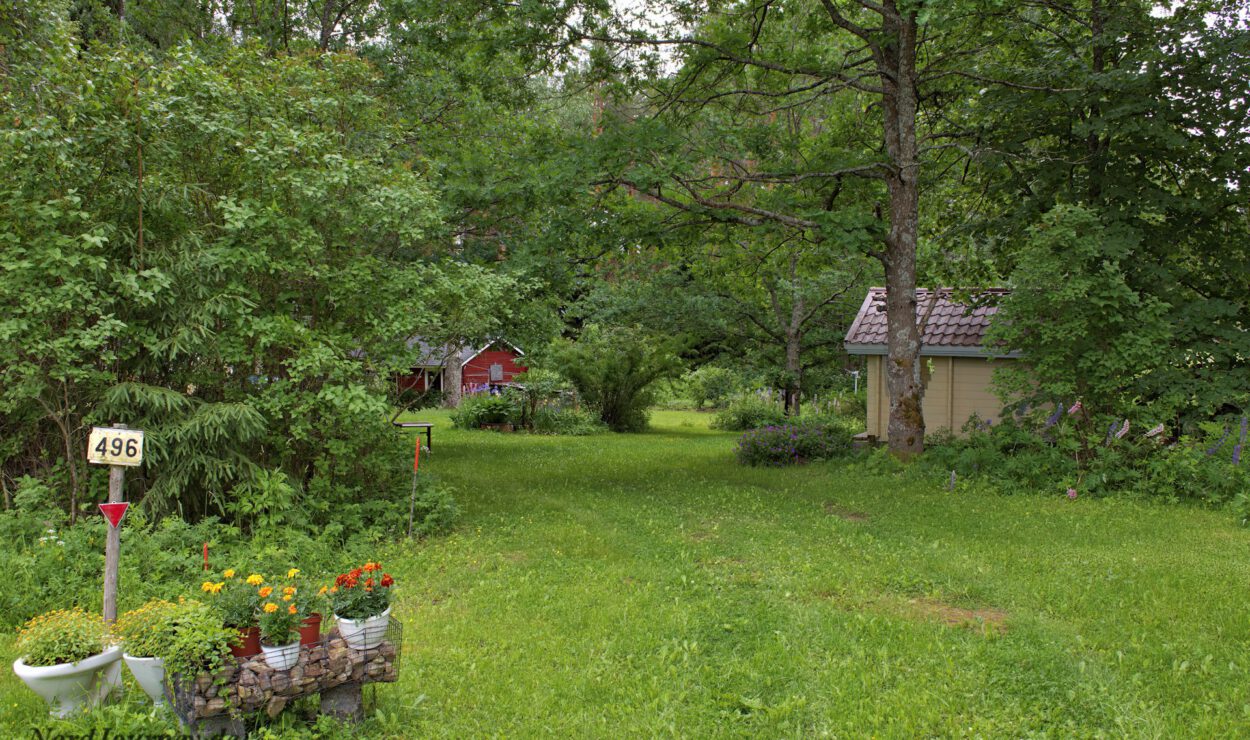 Ein üppiger grüner Garten mit kleinen Strukturen, umgeben von Bäumen. Im Vordergrund steht ein Steinpflanzgefäß mit Blumen und einem Schild mit der Aufschrift „496“.