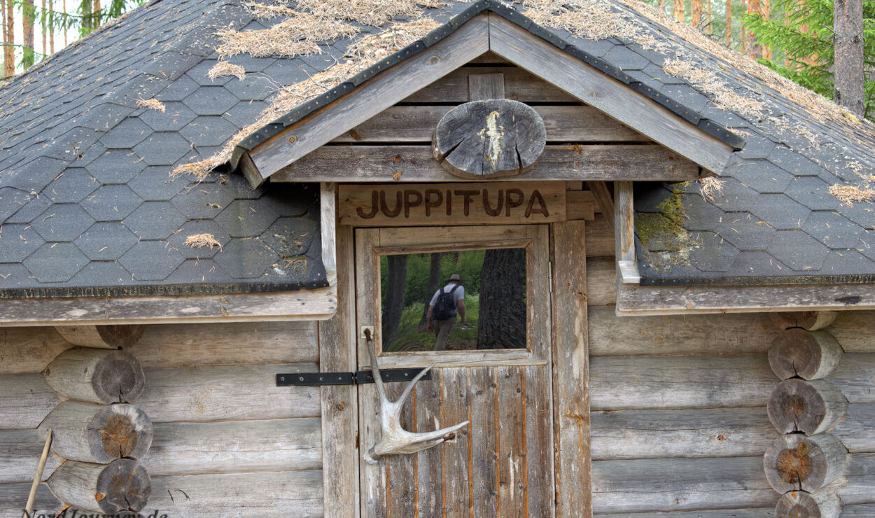 Holzhütte mit sechseckigem Schindeldach in einem Wald. Über der Tür, die mit Geweihdekor verziert ist, steht der Name „Juppitupa“. Durch ein kleines Fenster in der Tür ist die Waldlandschaft sichtbar.