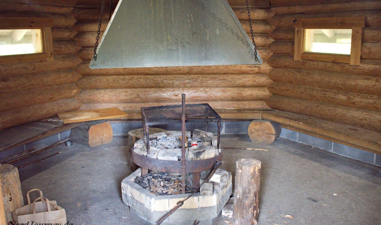 Innenansicht einer Blockhütte mit zentraler Feuerstelle, Steinsockel und Metallkamin. Kreisförmige Holzsitze umgeben den Bereich. Natürliches Licht fällt durch kleine Fenster ein.