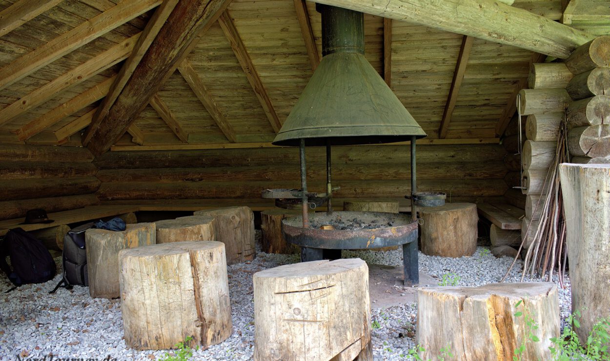 Ein rustikaler Unterstand aus Holzstämmen mit einer zentralen Feuerstelle aus Metall und mehreren groben Holzhockern auf einem Kiesboden. Das Dach besteht aus Holzbalken.