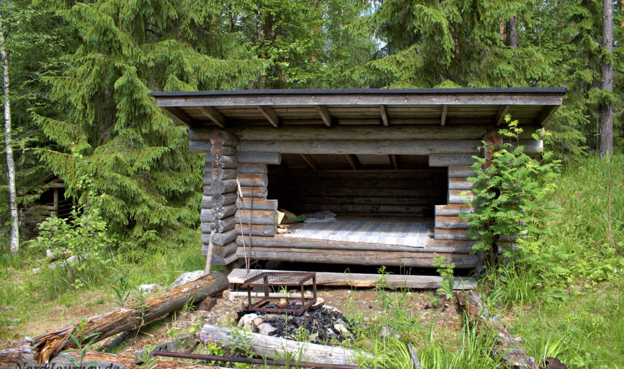 Ein kleiner Holzunterstand auf einer Waldlichtung mit einem einfachen Bett darin und einer Feuerstelle davor.