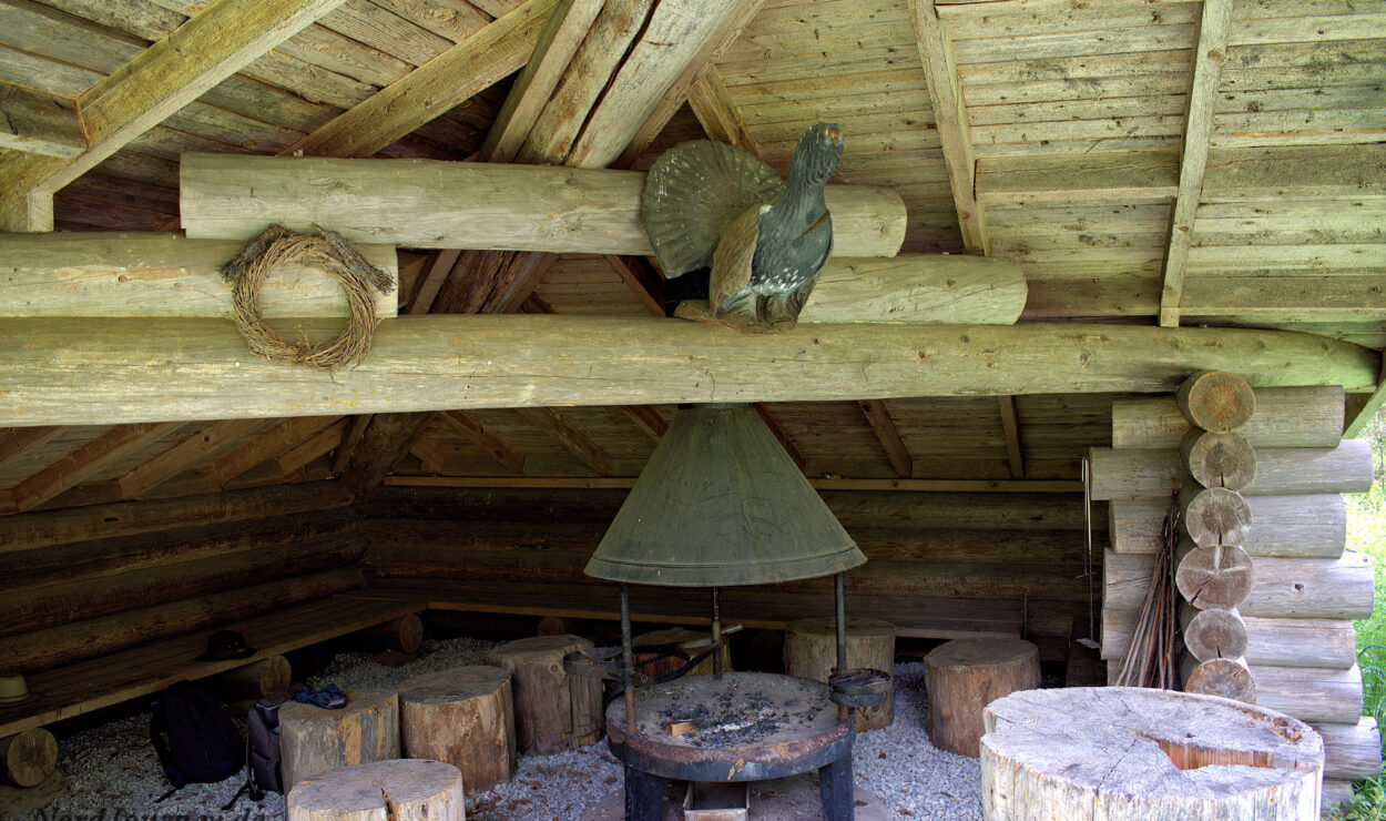 Rustikales Blockhaus-Interieur mit Holzbänken, einer zentralen Feuerstelle und einer darüber angebrachten dekorativen Vogelfigur.