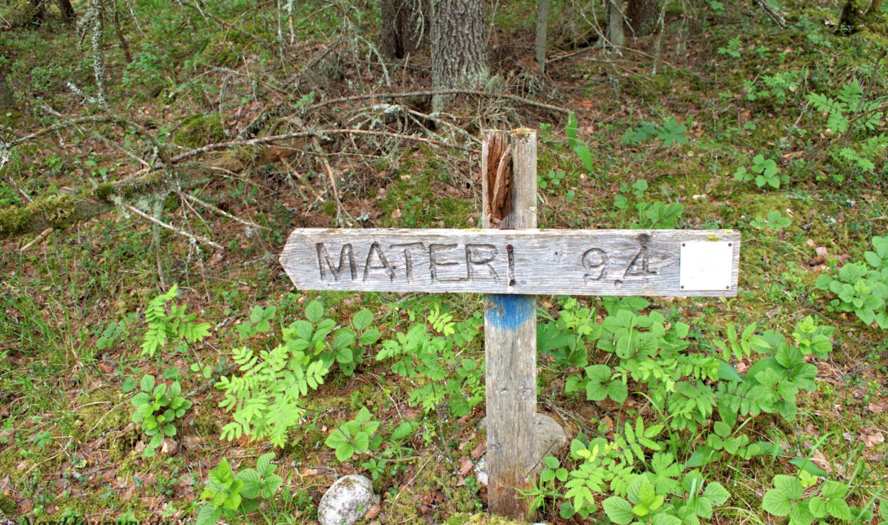 Hölzerner Wegweiser in einem Wald, der in Richtung „Materi“ zeigt, mit einer Entfernung von 9,4, umgeben von Grün und Bäumen.