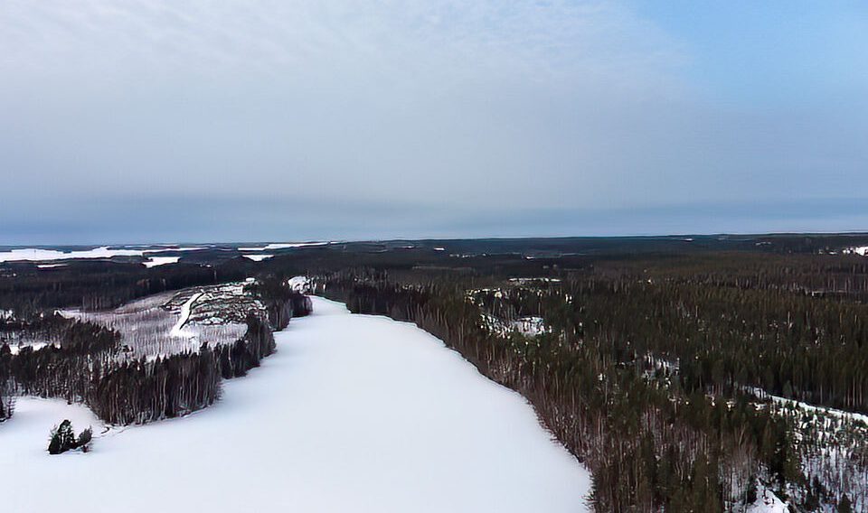 Luftaufnahme einer schneebedeckten Landschaft mit einem gefrorenen Fluss, umgeben von dichten Wäldern unter einem bewölkten Himmel.