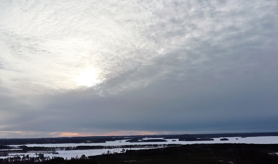 Luftaufnahme einer Winterlandschaft mit schneebedeckten Feldern und vereinzelten Bäumen unter einem bewölkten Himmel mit teilweise sichtbarer Sonne.