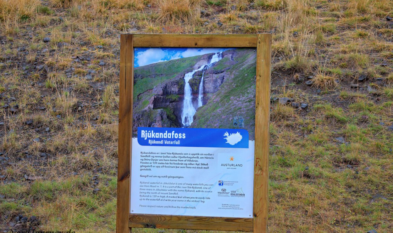 Vor einem grasbedeckten Hügel steht ein Holzschild mit Informationen zum Wasserfall Rjukandafoss.