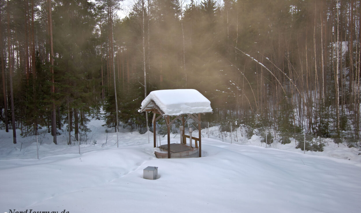 Ein schneebedeckter Brunnen steht auf einer verschneiten Waldlichtung, im Hintergrund sind hohe Bäume zu sehen.