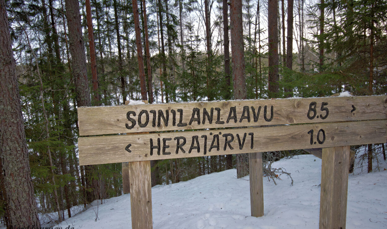 Holzschild im verschneiten Wald mit Wegbeschreibung: links nach Soinilanlaavu (8,5 km), rechts nach Herajärvi (1,0 km).