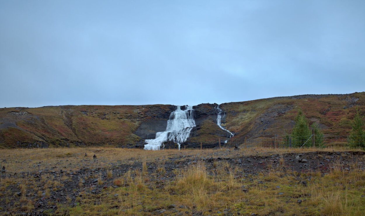 Ein Wasserfall, der einen felsigen Hügel hinunterstürzt, mit Gras im Vordergrund und einem bewölkten Himmel darüber.