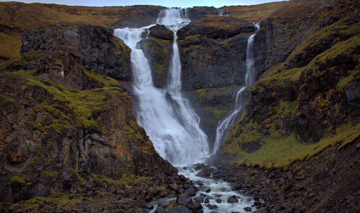 Wasserfall, der über felsige Klippen mit moosbedeckten Flecken herabstürzt, umgeben von zerklüftetem Gelände.