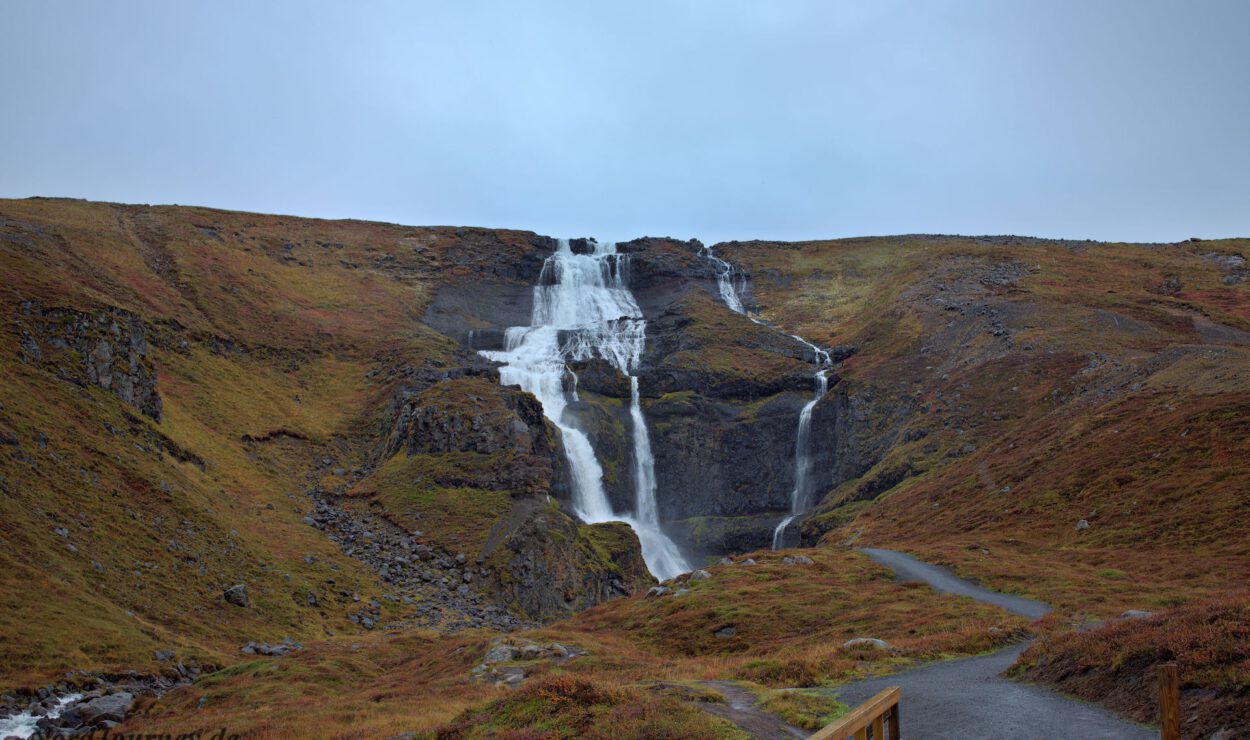 Ein Wasserfall stürzt eine mehrstufige Felsklippe hinab, umgeben von herbstlich gefärbter Vegetation unter einem bewölkten Himmel. Ein Pfad führt hinauf zum Aussichtsbereich im Vordergrund.