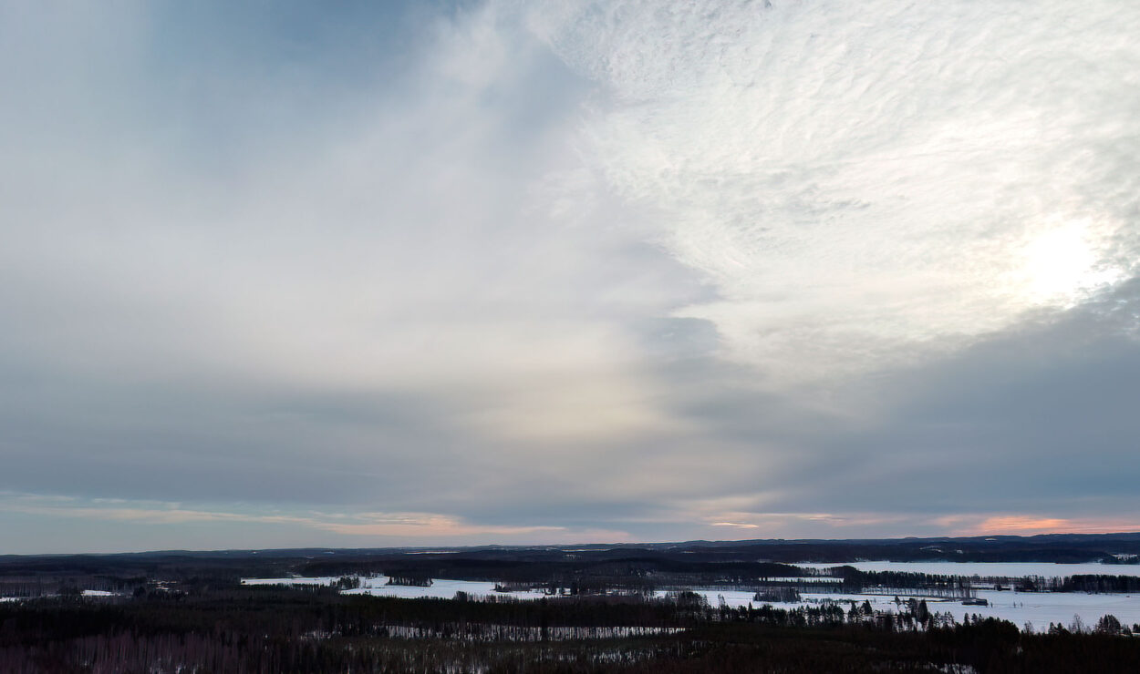 Eine weite Landschaft mit schneebedeckten Feldern, Wäldern und einem bewölkten Himmel, über dem die Sonne tief am Horizont steht.