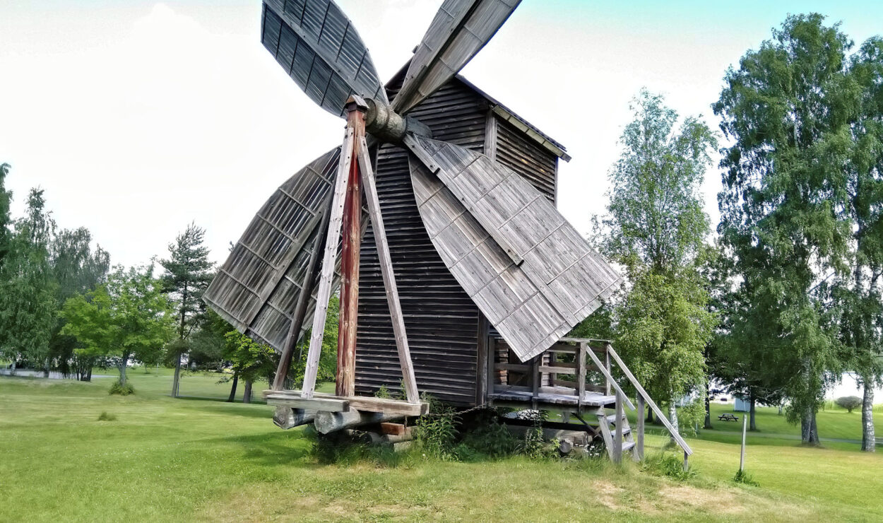 Eine alte hölzerne Windmühle mit großen Flügeln steht auf einer Wiese, umgeben von Bäumen unter einem klaren Himmel.