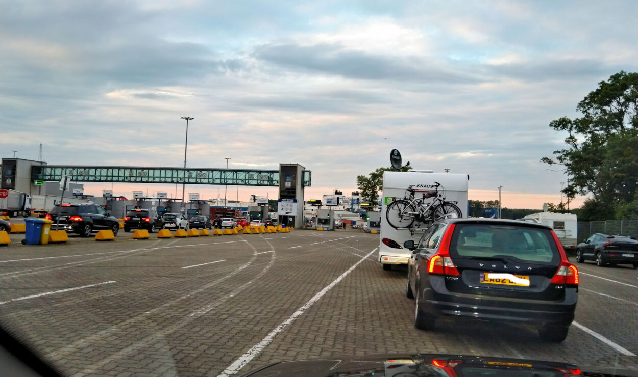 Fahrzeuge, darunter Autos und ein Wohnmobil mit Fahrrädern, stehen unter einem bewölkten Himmel in mehreren Spuren an einem Fährterminal aufgereiht. Im Hintergrund ist eine Fußgängerbrücke zu sehen.