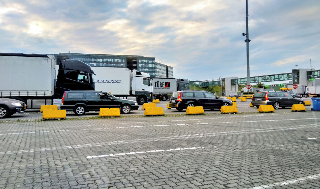 Lastwagen und Autos stehen aufgereiht auf einem gepflasterten Parkplatz mit gelben Betonbarrieren und Gebäuden im Hintergrund unter einem bewölkten Himmel.