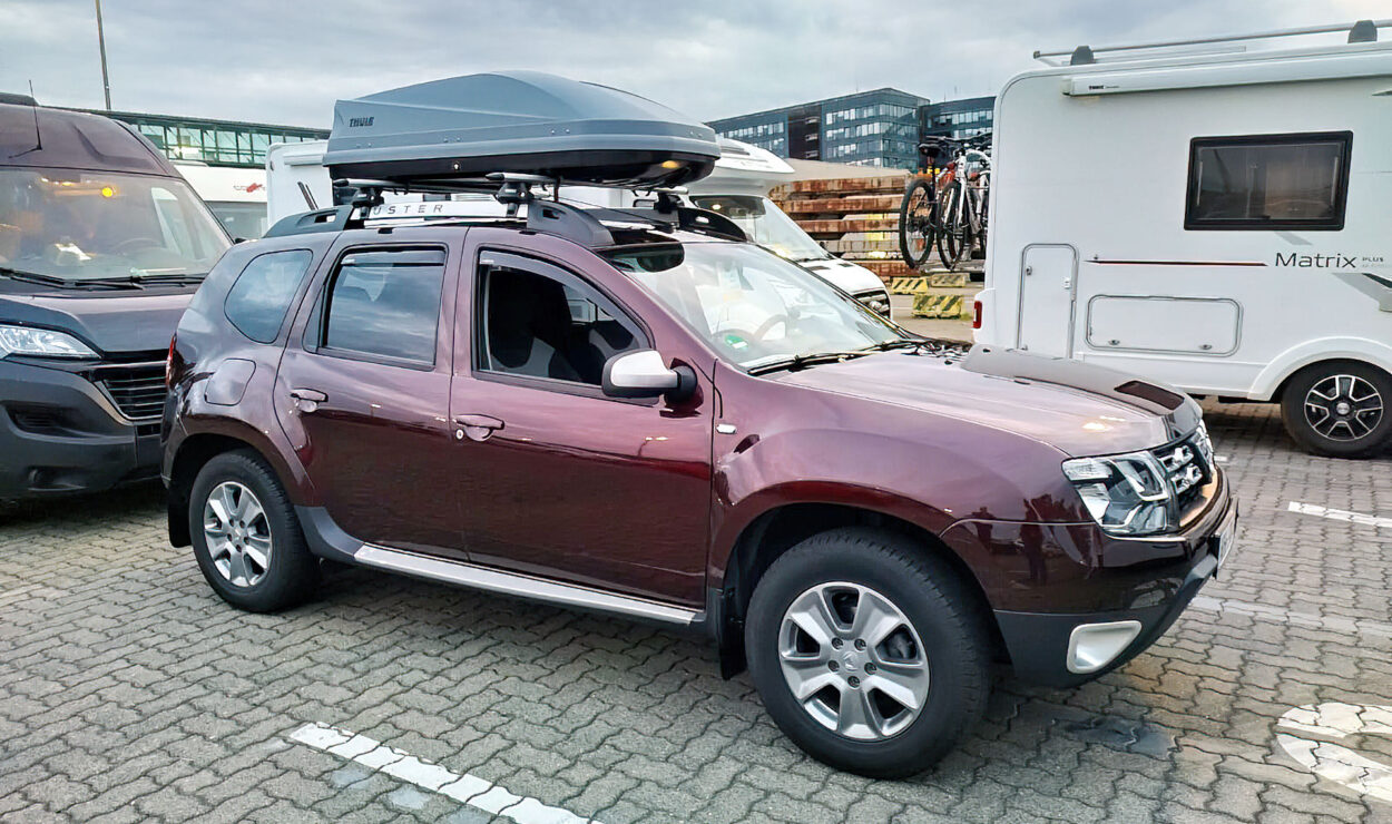 Kastanienbrauner SUV mit einer Dachbox, der auf einem Parkplatz neben einem Wohnmobil geparkt ist.