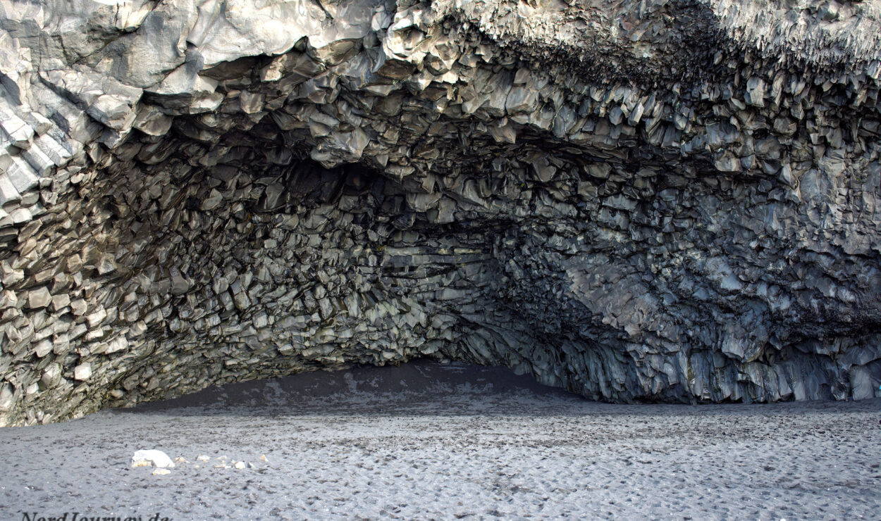 Höhle mit Basaltsäulen und dunklem Sandstrand, die einen symmetrischen Torbogen innerhalb einer felsigen Landschaft bilden.