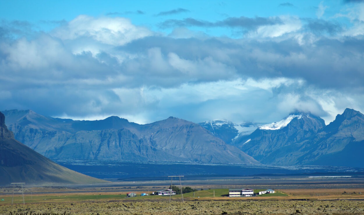 Ländliche Landschaft mit kleinen Gebäuden im Vordergrund, umgeben von weiten Ebenen. Majestätische Berge mit schneebedeckten Gipfeln unter einem wolkig blauen Himmel im Hintergrund.