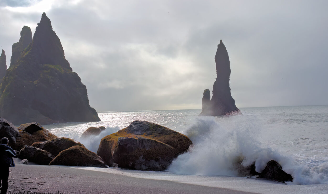 Wellen schlagen gegen felsige Felsnadeln an einem nebligen Strand mit Klippen in der Ferne und einem bewölkten Himmel.