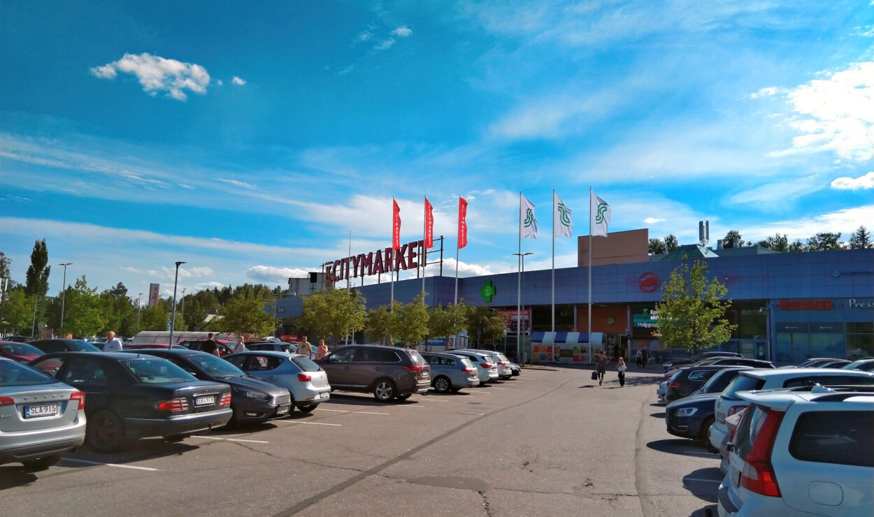 Parkplatz eines Einkaufszentrums mit geparkten Autos an einem sonnigen Tag; auf dem Gebäude sind Fahnen und das Wort „CITYMARKET“ zu sehen.