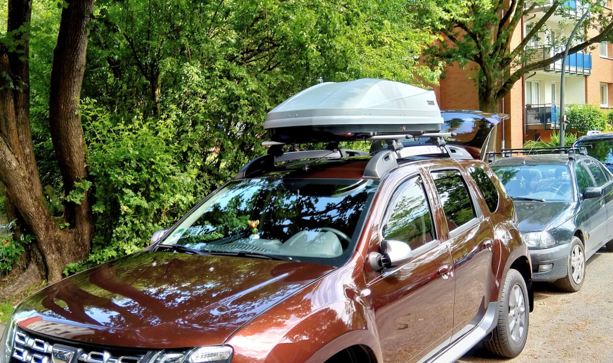 Brauner SUV mit Dachbox, geparkt auf einer begrünten Straße neben einem anderen Auto. Im Hintergrund Bäume und ein Gebäude.