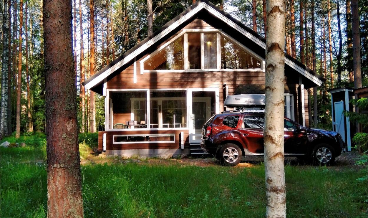 Eine Holzhütte mit großen Fenstern liegt eingebettet zwischen hohen Bäumen. Davor parkt ein roter SUV. Sonnenlicht fällt durch den Wald.