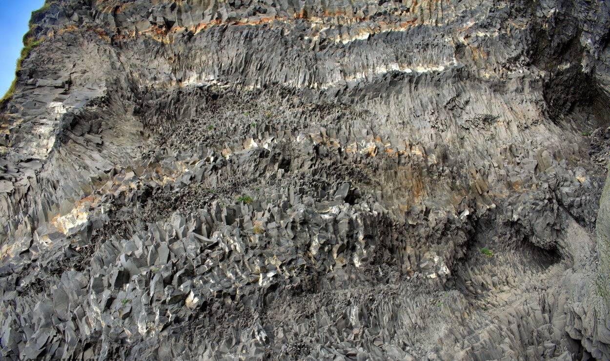 Nahaufnahme einer felsigen Klippe mit zerklüfteten, geschichteten Formationen in verschiedenen Grautönen und einem Hauch grüner Vegetation.