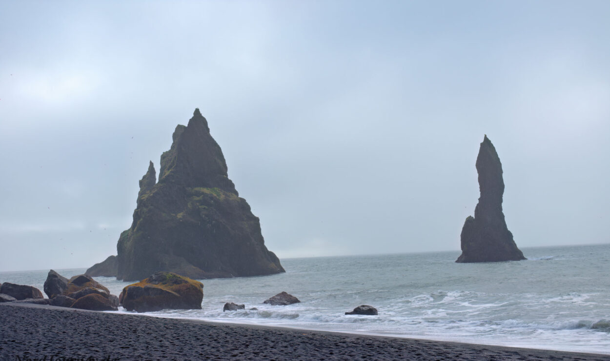 Zwei hohe Felssäulen aus Basalt stehen nahe der Küste eines grauen, bewölkten Strandes, an dessen schwarzen Sand die Wellen sanft brechen.